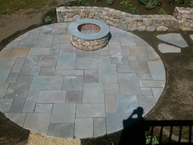 Paved stone patio with a circular fire pit and stone retaining wall in a backyard garden.