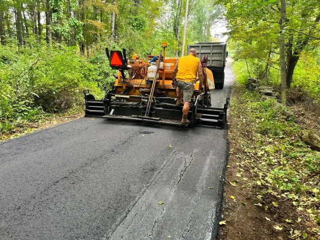 Road paving machine laying asphalt on a road, worker in yellow shirt directing. Trees in background.