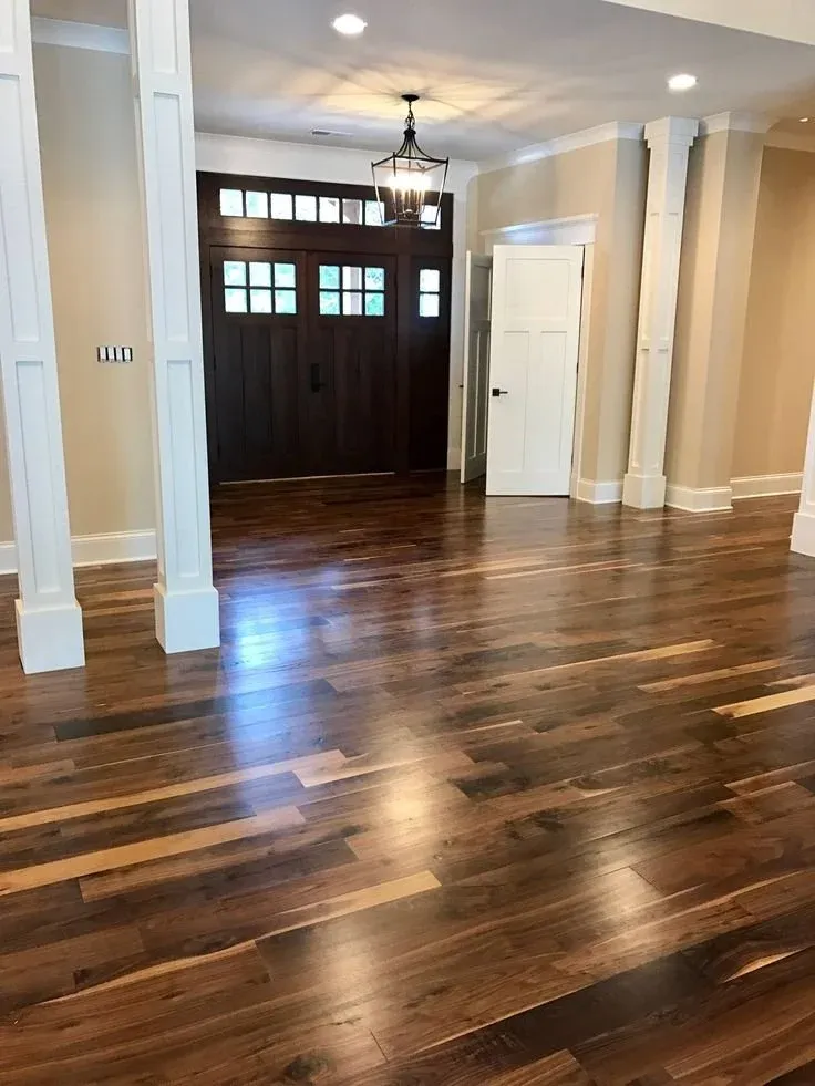 Interior of a house with dark wooden front door, hardwood floors, and beige walls. Ceiling light fixture and natural light from windows above the door.