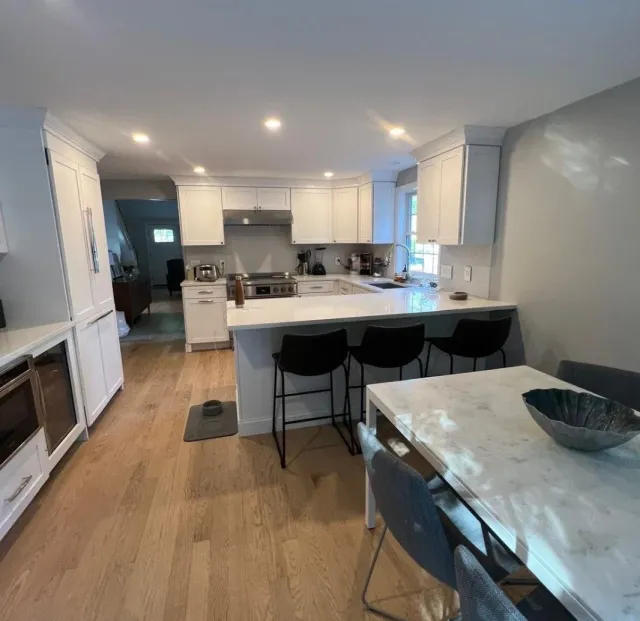Modern kitchen with white cabinets, stainless steel appliances, a marble island, wooden flooring, black bar stools, and a dining table with gray chairs.