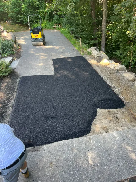 Asphalt being laid on a driveway, worker nearby with a compactor in the background, greenery in setting.