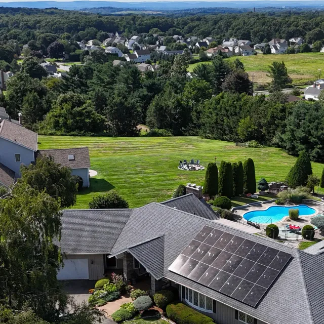 Aerial view of a suburban neighborhood with a house featuring solar panels, a manicured lawn, and a backyard swimming pool. The surrounding area has a variety of homes, trees, and open green spaces, with a scenic landscape in the background.