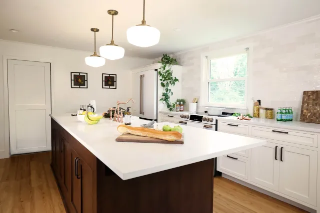 Modern kitchen with white cabinets, a large island with a white countertop, hanging pendant lights, a window, and various kitchen items including a loaf of bread, bananas, green apples, and bottles of water.