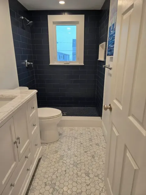 Bathroom with white vanity, toilet, and black tiled shower area with a window, white hexagon tile floor, and a white door.