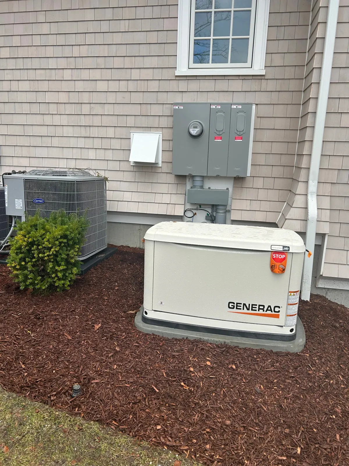 Outdoor view of a house's electrical equipment including a Generac generator, an air conditioning unit, electrical meter, and electrical panels on the beige house siding, with red mulch ground cover and a small shrub.