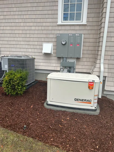 Outdoor view of a house's electrical equipment including a Generac generator, an air conditioning unit, electrical meter, and electrical panels on the beige house siding, with red mulch ground cover and a small shrub.