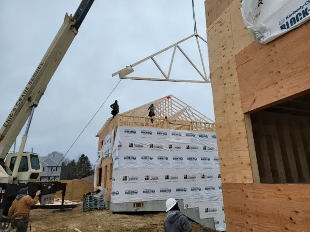 Crane lifting a wooden roof truss onto a partially built house at a construction site. Cloudy day.
