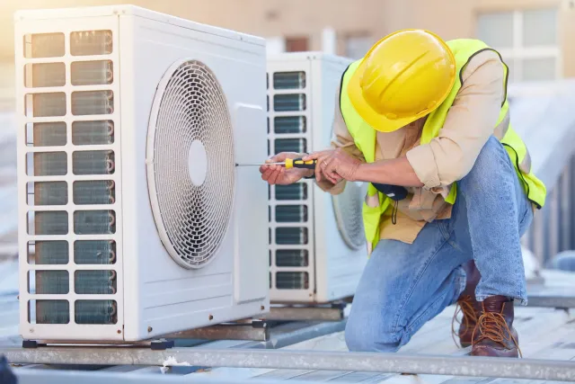 A technician in a yellow hard hat and safety vest uses a screwdriver to repair an outdoor air conditioning unit.