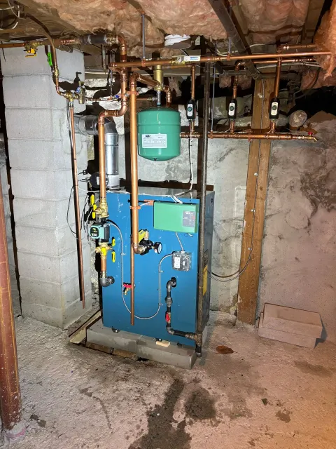 Blue boiler with copper pipes in a basement. Green expansion tank, concrete walls, and dirt floor.
