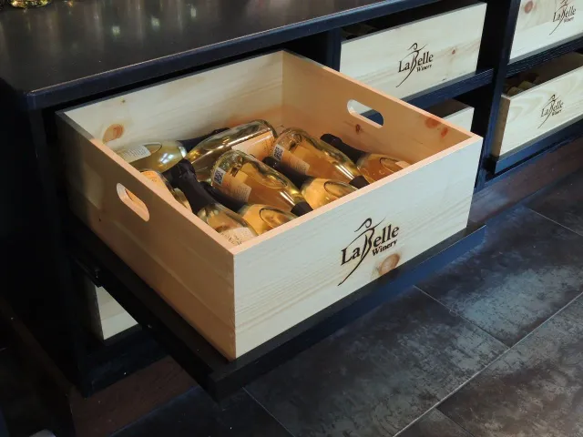 A wooden crate labeled La Belle Winery contains several bottles of white wine with gold foil tops, placed on a black shelf in a wine store.