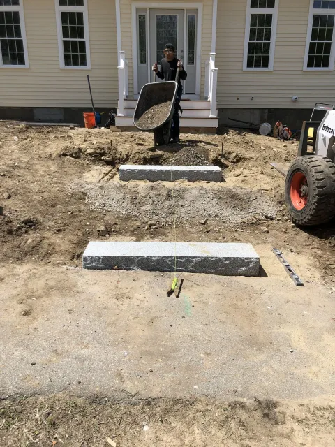Construction of steps leading to a house. A worker dumps gravel. Bobcat on the right.