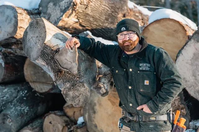 A man with glasses and a beard wearing a black beanie and a black work jacket with the logo 'Taylor Tree Service' on it, stands next to cut logs with snow on top in an outdoor, winter setting.