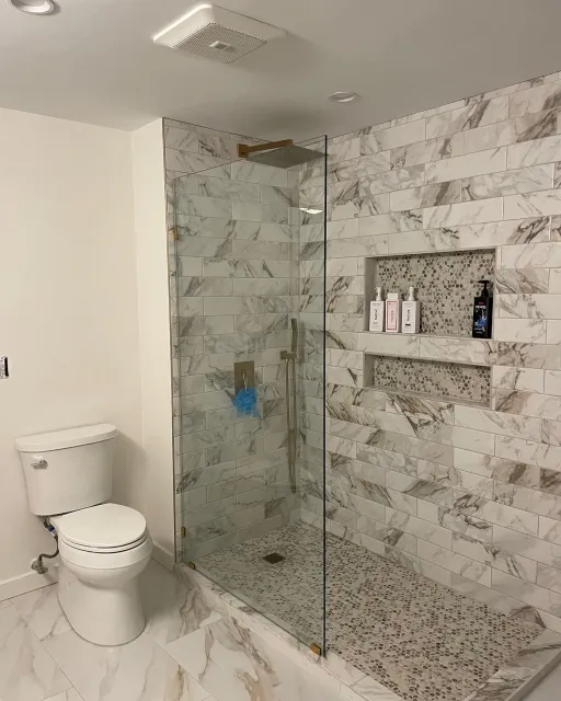 A modern bathroom with a glass-enclosed shower featuring marble-look tile, pebble flooring, and built-in shelving.