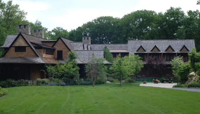 A large house with a grassy front yard, surrounded by trees and bushes, and a gravel driveway with a golf cart and wheelbarrow.