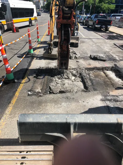 A construction site on a city street shows an excavator digging into the asphalt, creating a large hole. Orange traffic cones and barriers surround the work area, with a bus and parked cars visible in the background.