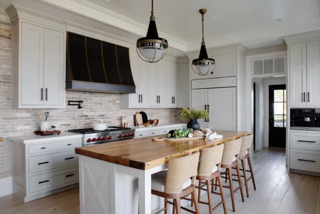 Hardwood floor installation in a Marshfield kitchen with white cabinets, a brick backsplash, a wooden island with seating, a black and brass stove hood, and two black pendant lights.