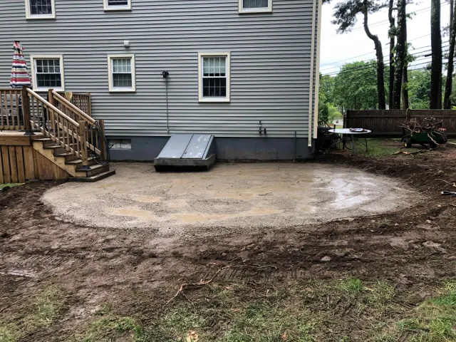 Backyard with a freshly leveled patio area next to a house, wooden deck, and grassy yard. Large metal cellar door, outdoor table with chairs, and a wheelbarrow with twigs are visible.