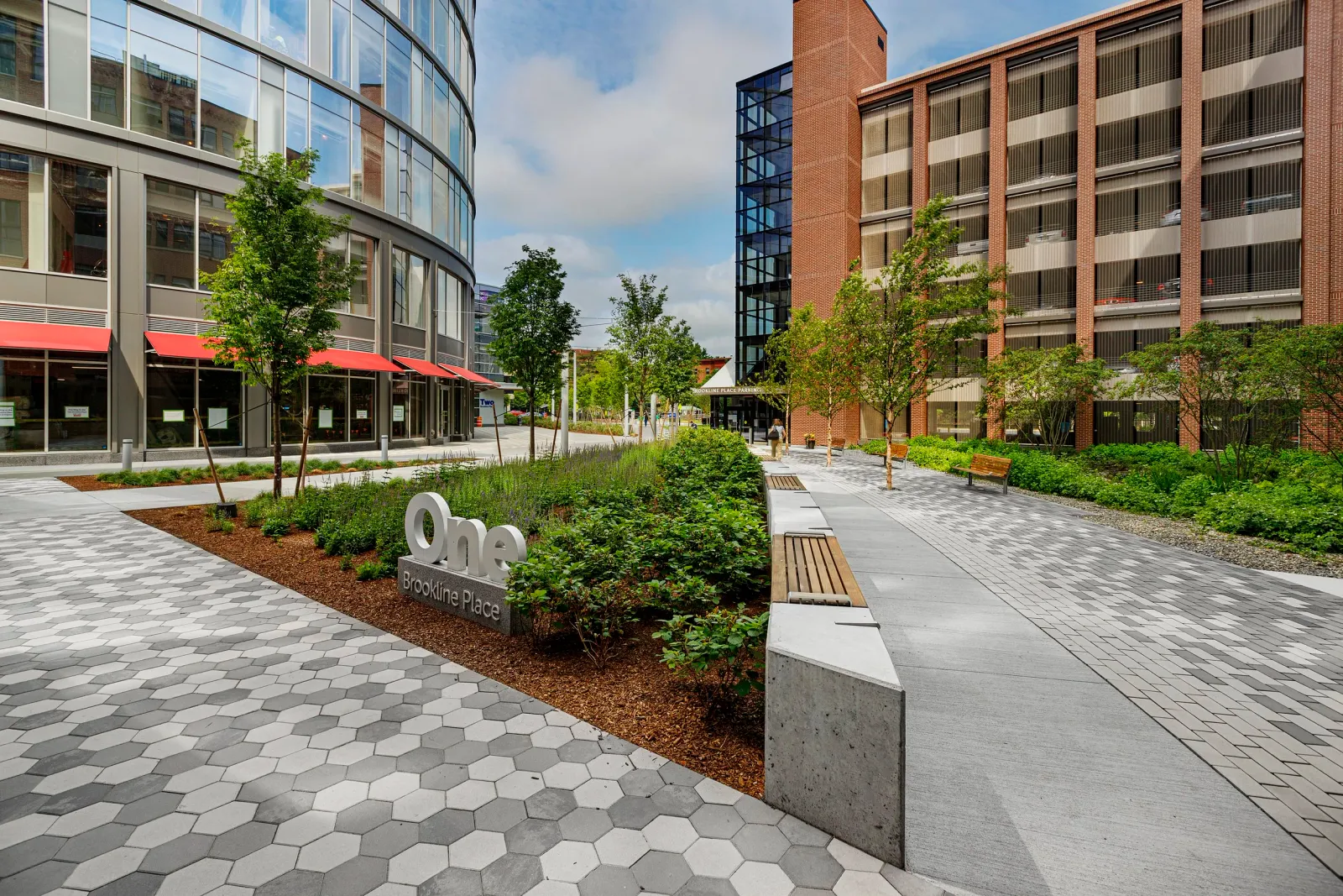 Modern outdoor urban plaza with trees, benches, and a mix of hexagonal and rectangular paving stones, surrounded by contemporary glass and brick buildings.