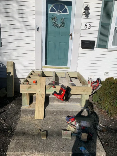 Front porch under construction with wooden frame, tools, and supplies on the steps and ground in front of a house with white siding, teal door with wreath, and black mailbox.