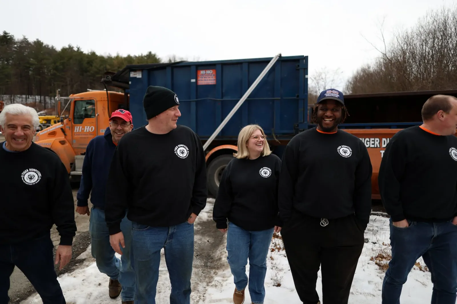 Group of six people walking outdoors in winter, wearing black sweatshirts with a logo, smiling and talking, with snow on the ground and trucks in the background.
