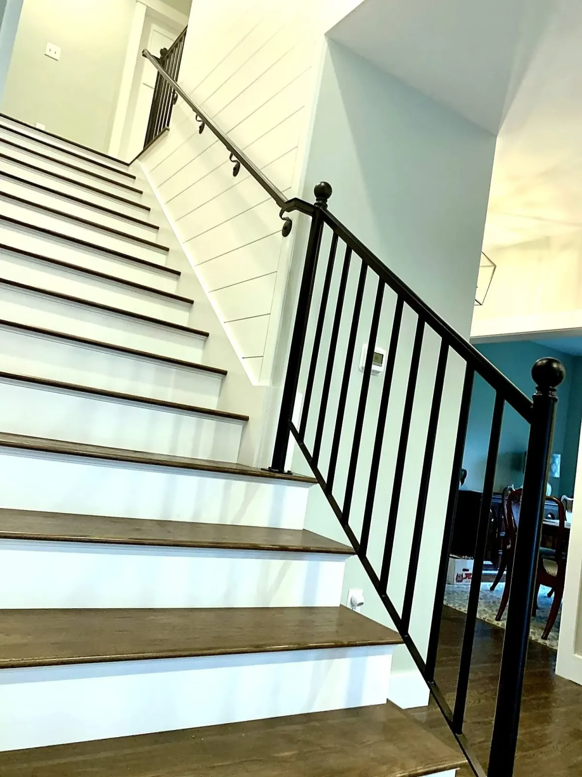 Inside view of a staircase with wooden steps, black metal railing, white wainscoting, and a glimpse of the living room area with chairs, a table, and a box in the background.
