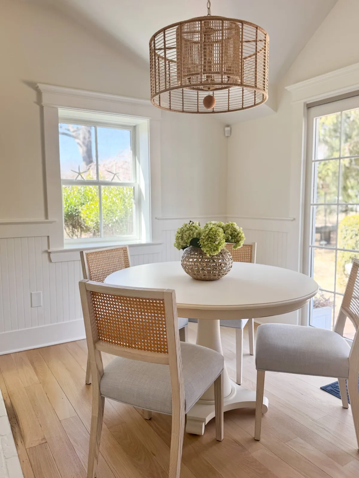 Hardwood floor refinishing in a Pembroke dining room with a white round table, light-fabric chairs, woven basket vase of green and white flowers, large windows, and a wicker ceiling light fixture.