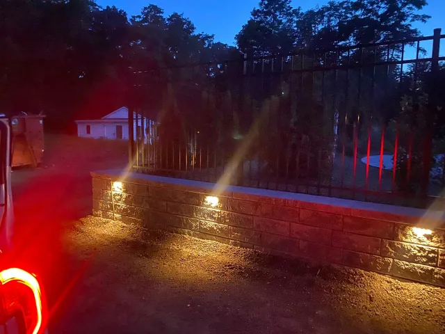 Low-angle view of a stone wall at twilight, featuring three warm LED lights illuminating the wall and surrounding ground.