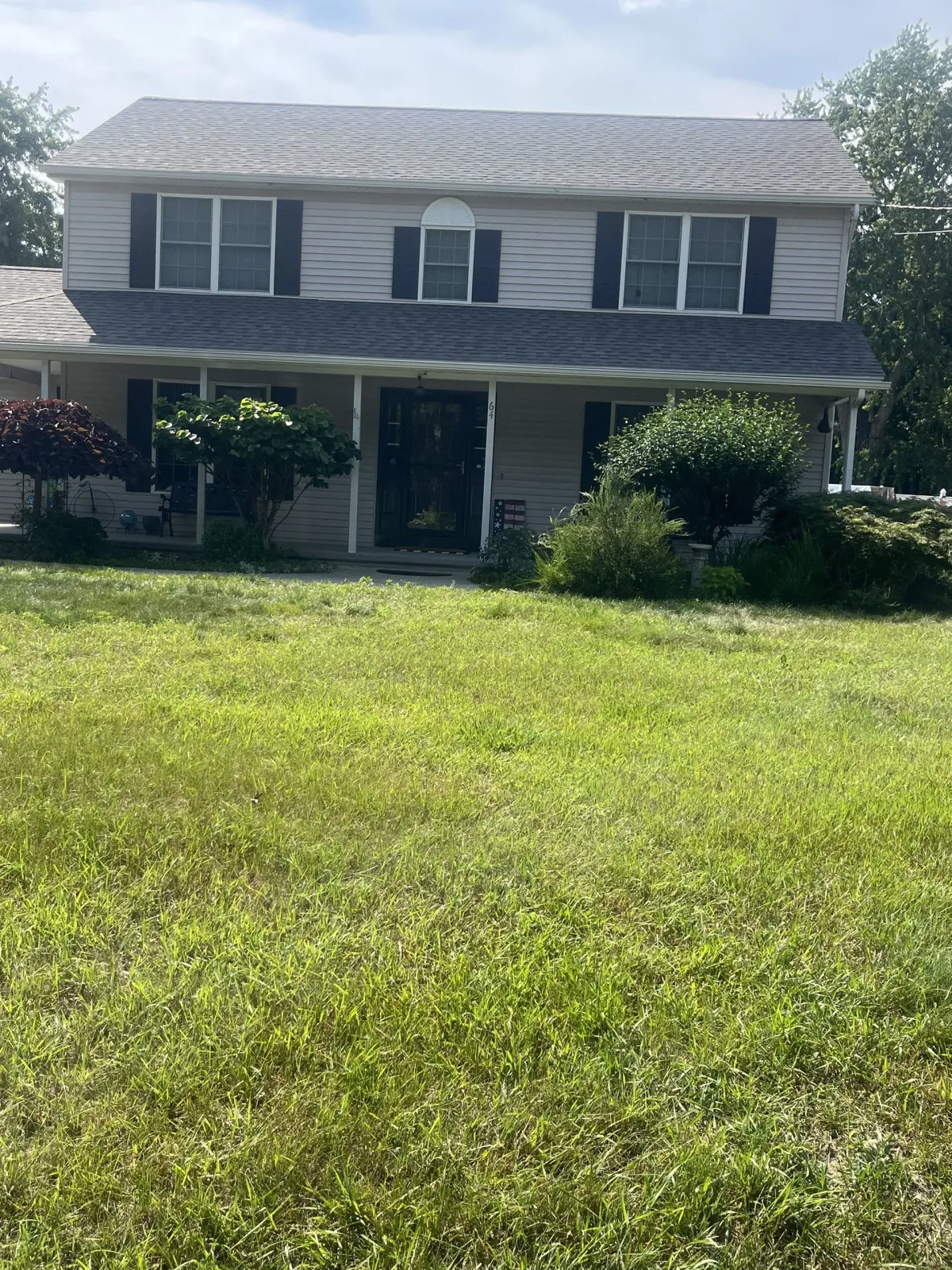 Two-story house with overgrown lawn; light gray siding, black shutters, shrubs, and a cloudy sky.