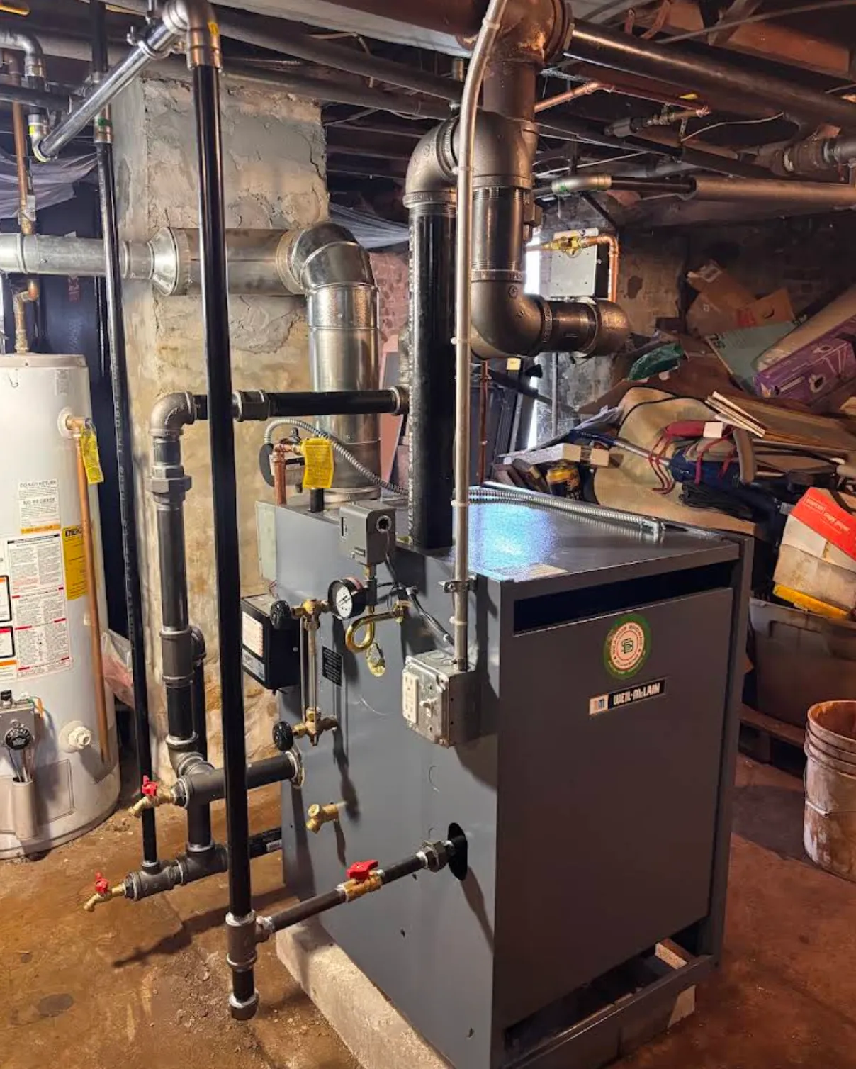 An industrial boiler system with black and gray pipes, a water heater on the left, and various gauges and valves, installed in a basement or utility room with unfinished walls and cluttered storage.