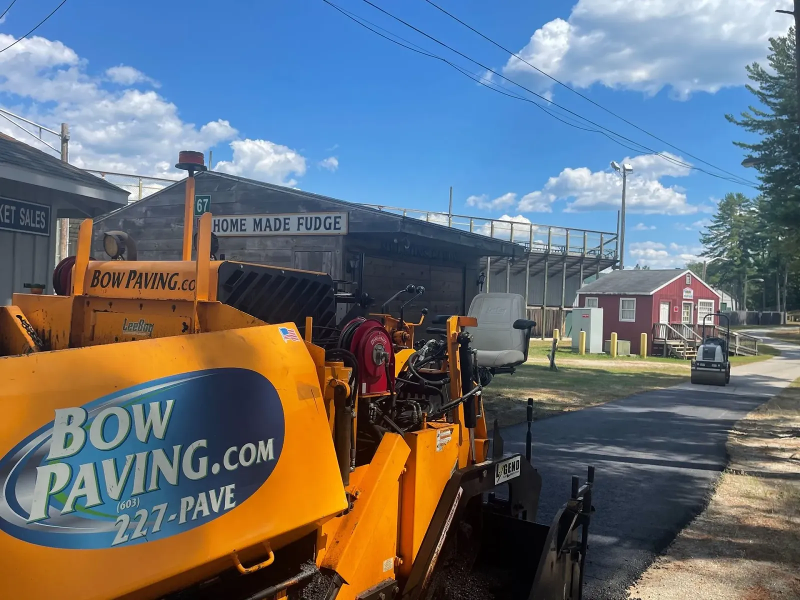 A yellow Bow Paving construction vehicle is parked on an asphalt path in front of a red building and stadium bleachers.
