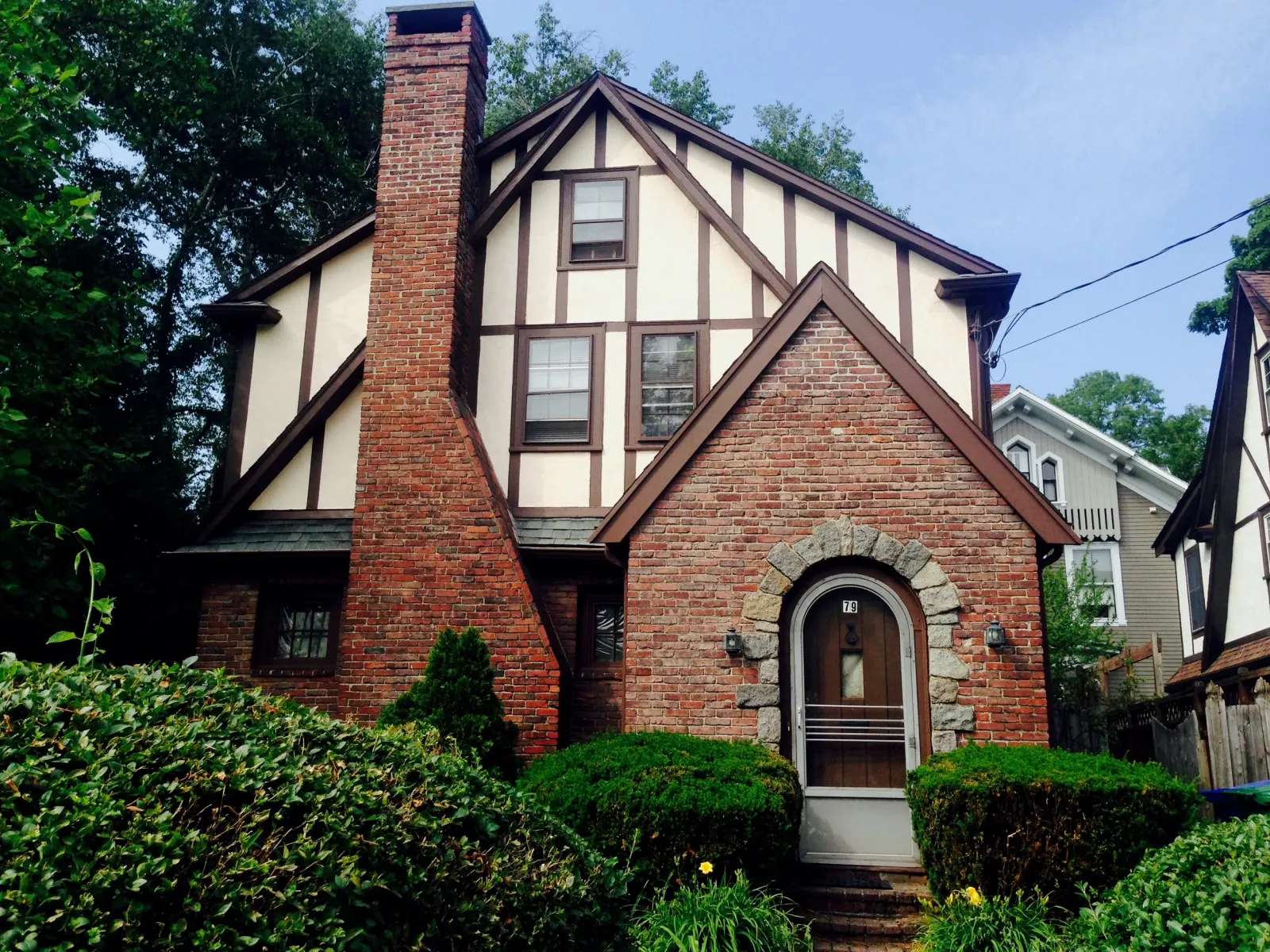 Brick and timber Tudor-style house with large chimney, arched door, and front garden in a residential neighborhood.