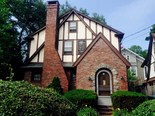 Brick and timber Tudor-style house with large chimney, arched door, and front garden in a residential neighborhood.