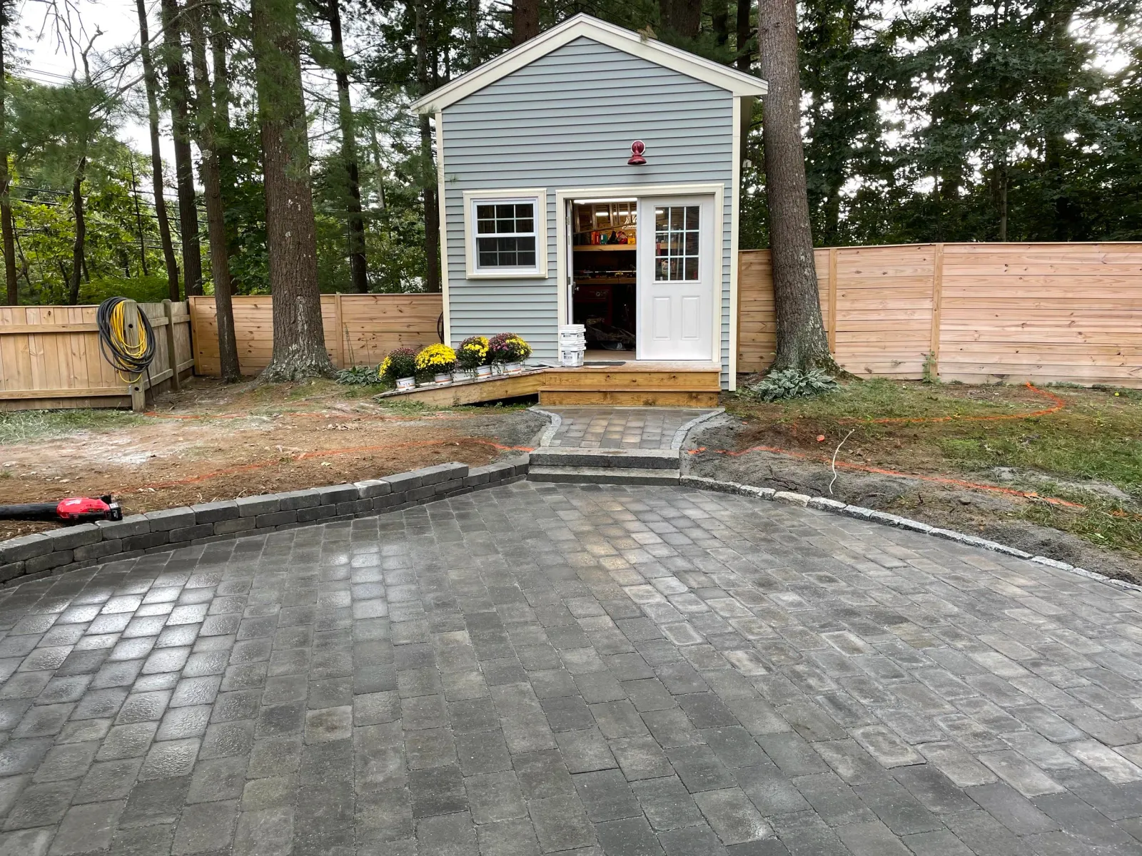 Backyard with paved patio, small shed, wooden fence, trees, and garden tools