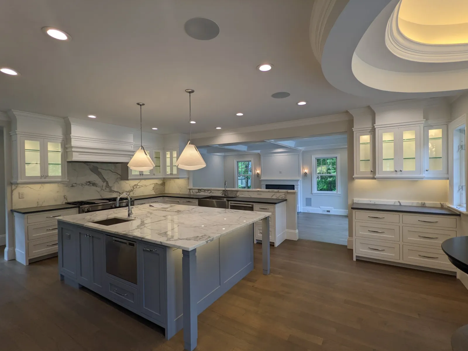 Modern kitchen with white cabinets, marble countertops, a central island with a sink and oven, pendant lighting, and wood flooring.