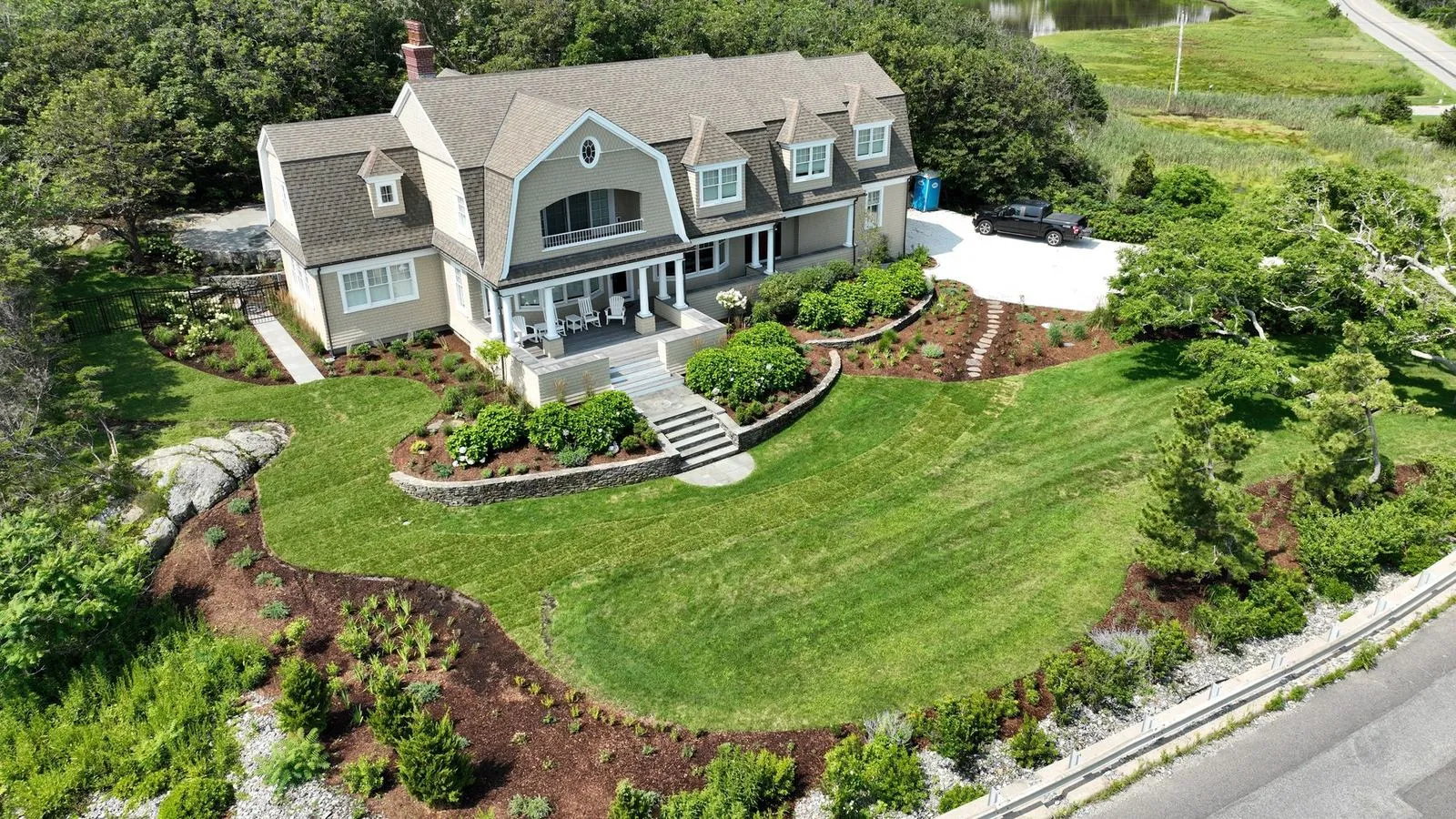 An aerial view of a shingle-style Cape Cod estate — manicured lawn, layered planting beds, stone retaining walls, and a coastal road in the distance