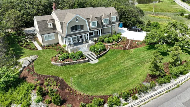 An aerial view of a shingle-style Cape Cod estate — manicured lawn, layered planting beds, stone retaining walls, and a coastal road in the distance