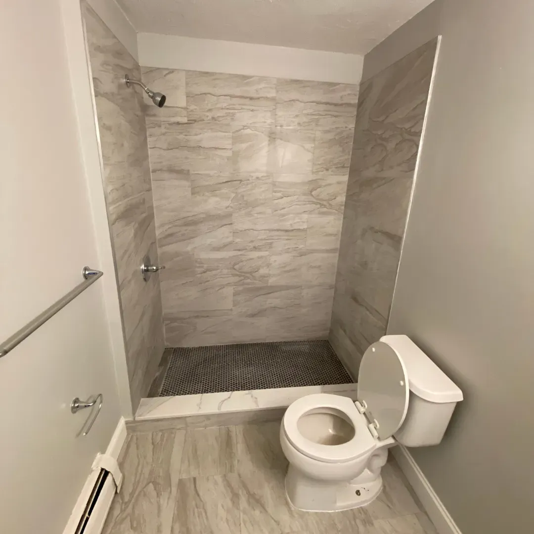 Stand-up shower with veined wood-look stone tile and white toilet by SF Construction in Marlborough, MA