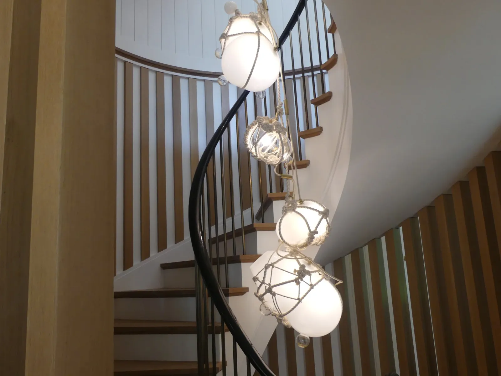 Interior view of a spiral staircase with wooden steps and black railing, featuring hanging white globe pendant lights wrapped in rope for decorative effect.