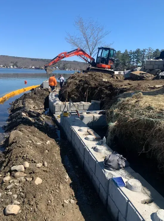 Construction crew building a shoreline retaining wall beside a lake, with an orange excavator on the bank.