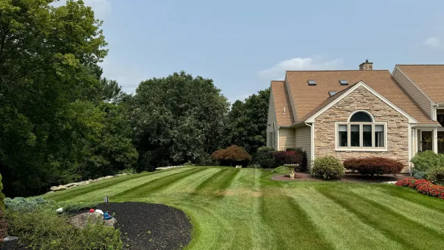 Lawn with striped grass in front of a stone house with a brown roof and a blue sky.