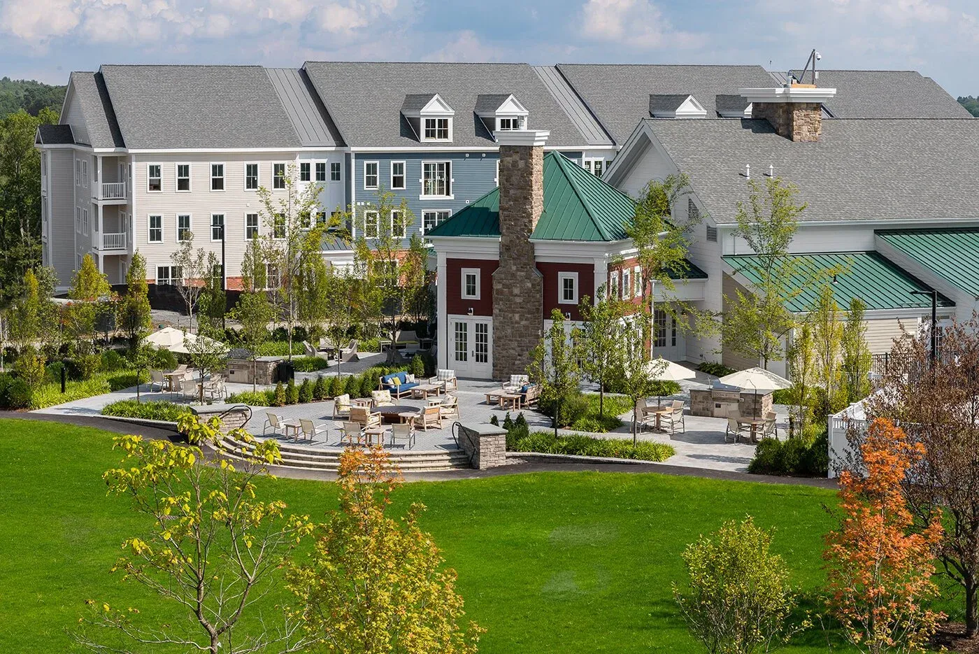 A commercial project of landscaped residential complex with a spacious functional patio area, surrounded by manicured green grass and trees, and multistory buildings with gray roofs and white, red, and blue exteriors.