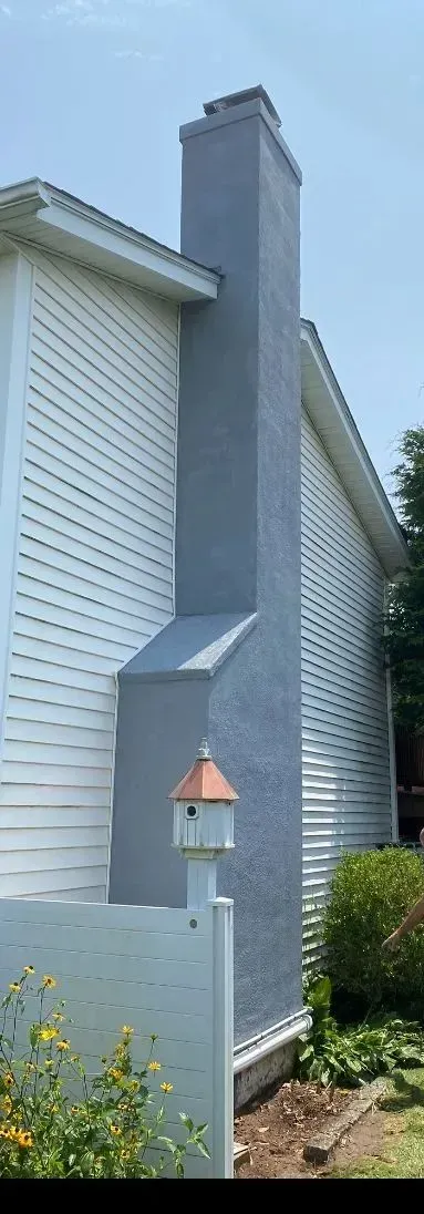 A tall, grey stucco chimney sits against the side of a white-sided house with a birdhouse in the foreground.