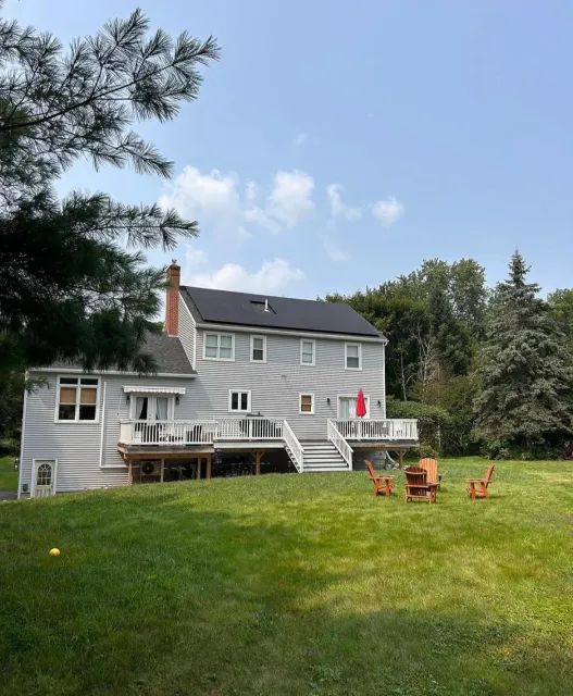 Backyard view of a two-story house with a white exterior, deck, and patio seating on a grassy lawn, surrounded by trees on a clear day.