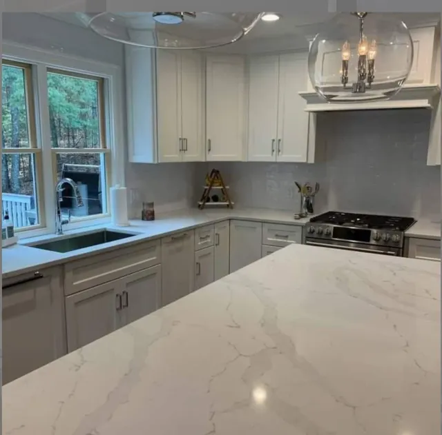Modern kitchen with white cabinets, marble countertop, stainless steel stove, and a window overlooking trees.