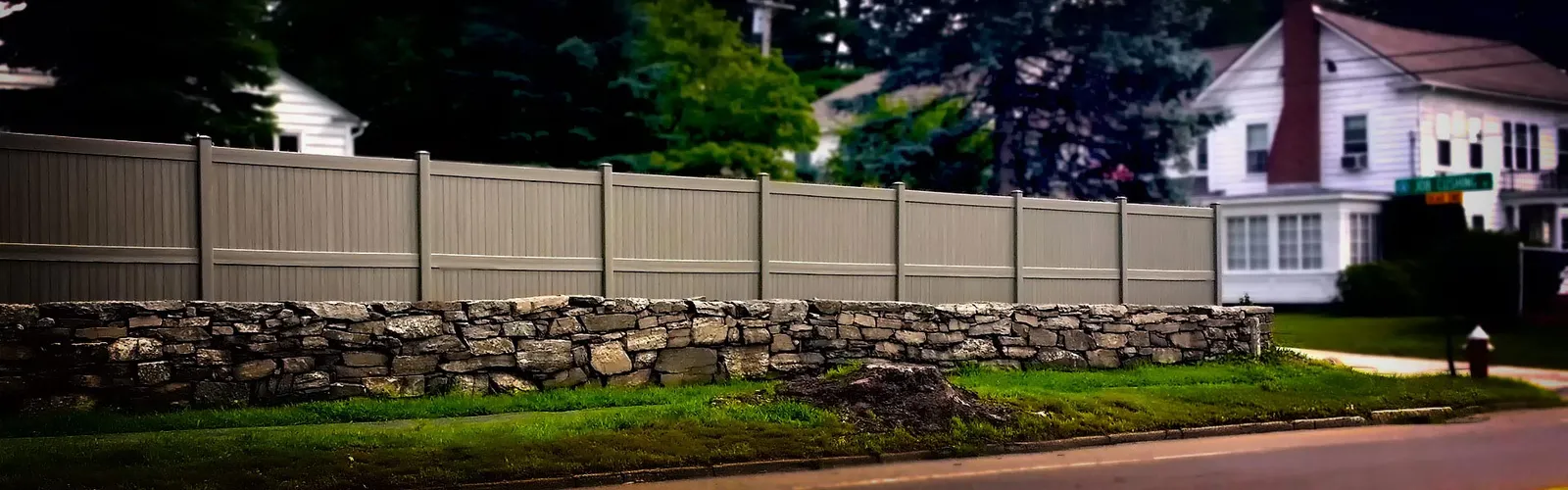 A light brown vinyl fence above stone wall along a residential street with a white house in the background