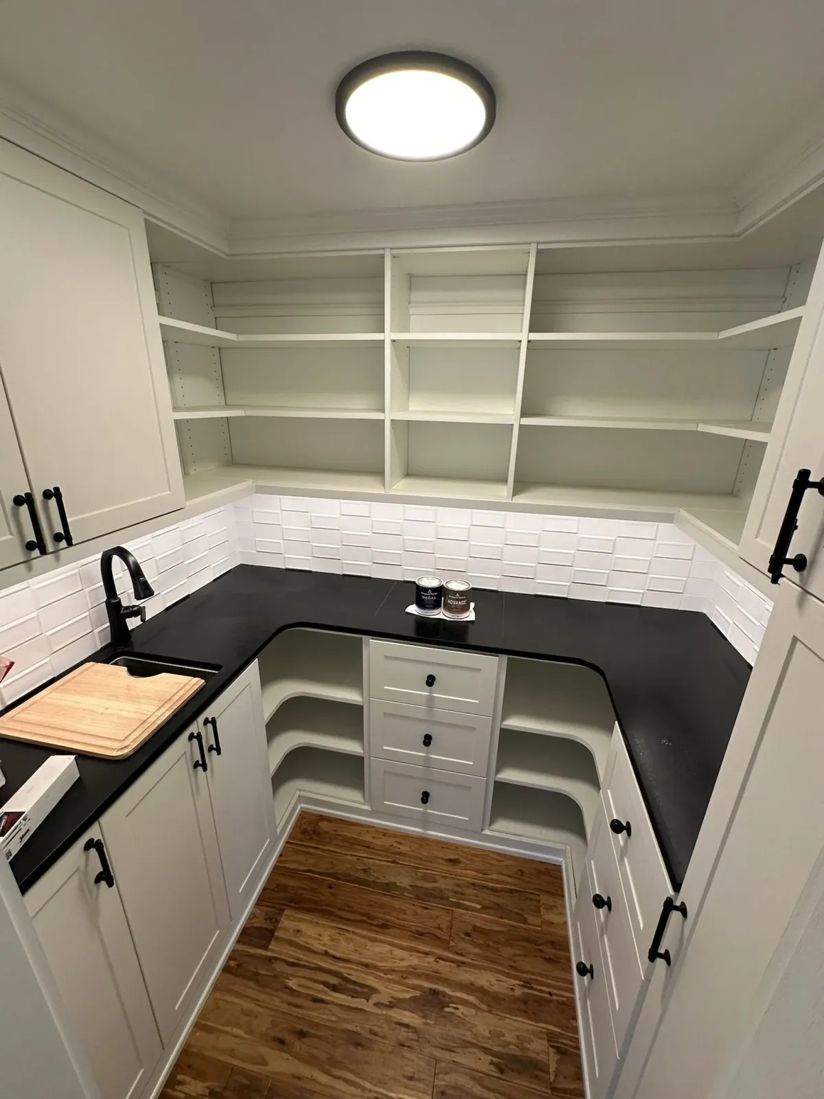 Empty kitchen with white cabinets, black handles, black countertop, white subway tile backsplash, black faucet, cutting board on counter, open shelving above, and hardwood floor.