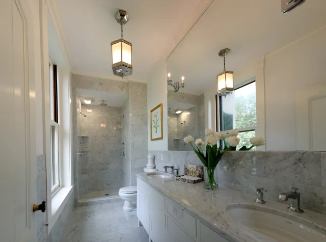 Bright bathroom with marble countertops, two sinks, a large mirror, a vase of white flowers, a window, a shower with multiple showerheads, and classic lighting fixtures.