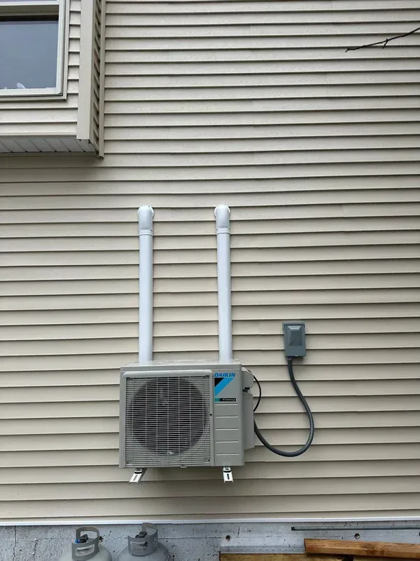 A Daikin ductless HVAC outdoor unit mounted on beige vinyl siding, with two vertical white conduits and a disconnect box.