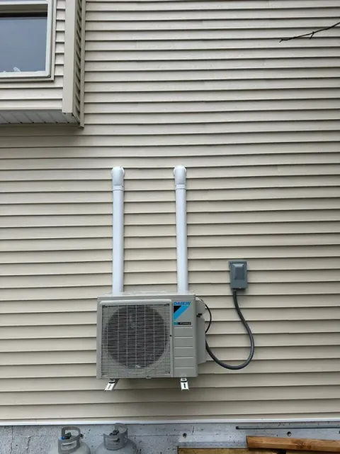 A Daikin ductless HVAC outdoor unit mounted on beige vinyl siding, with two vertical white conduits and a disconnect box.