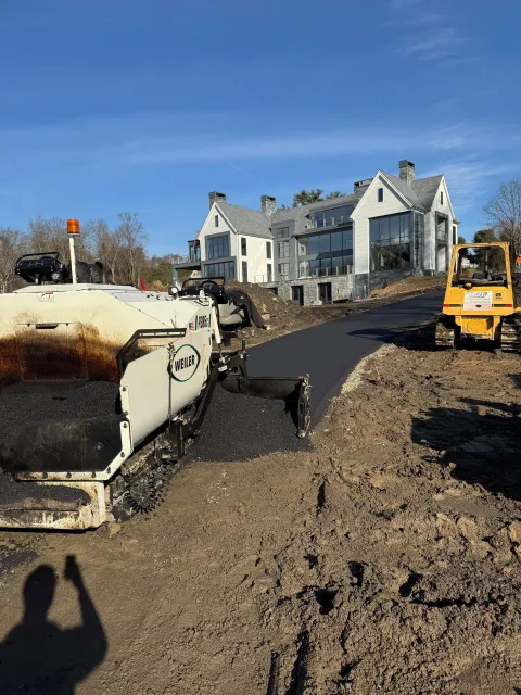Road paving equipment at a muddy construction site beside a large house under a blue sky
